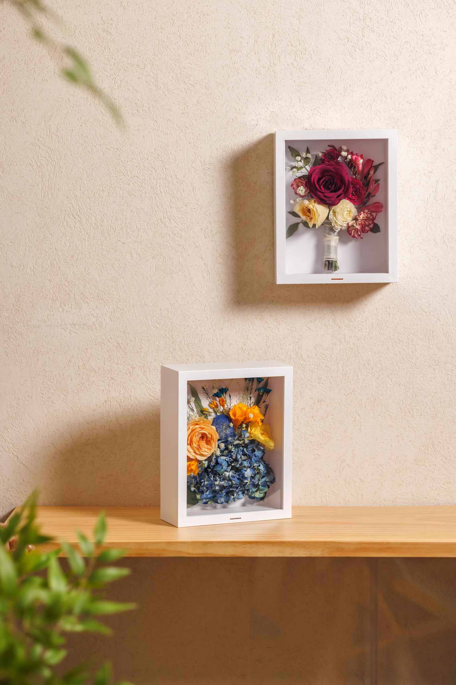 Two Wisedry shadow boxes displaying preserved bouquets: a red and cream rose arrangement hung on a wall, and a peach rose and blue hydrangea arrangement standing on a wooden shelf.